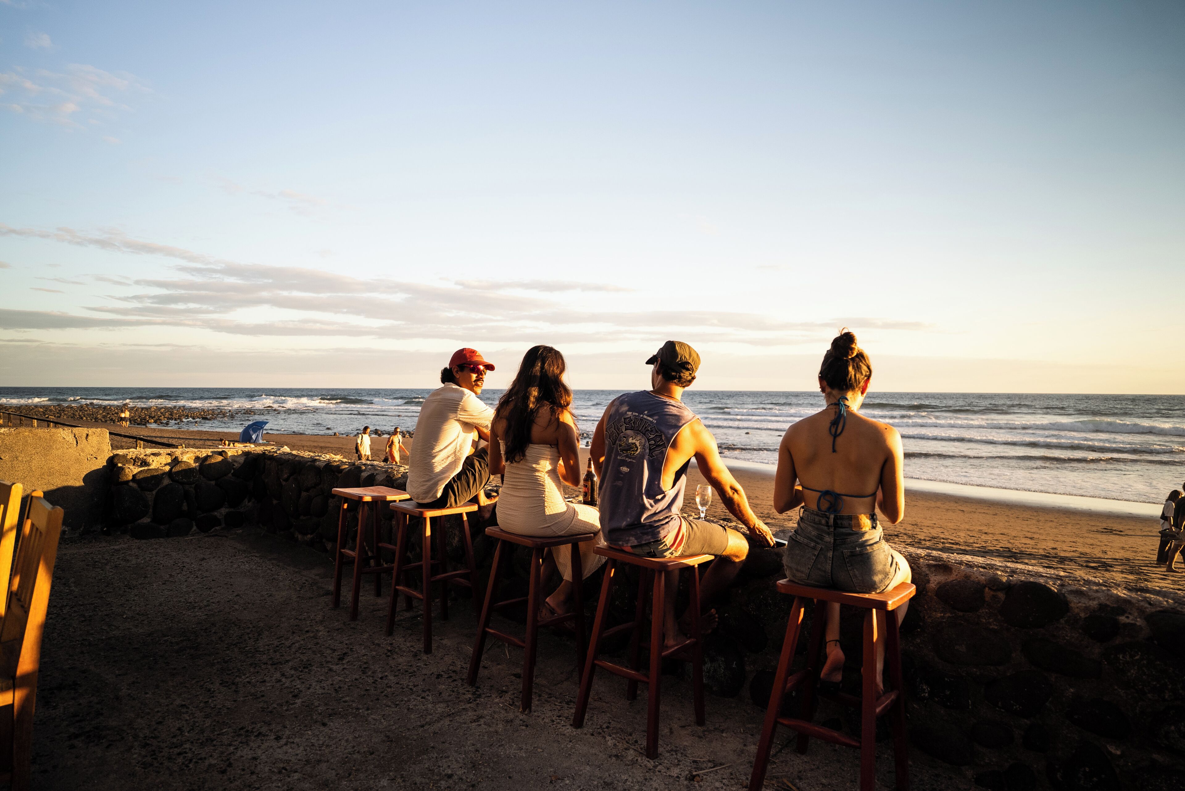 Un grupo de personas sentadas en taburetes en un bar frente a la playa, con vista al océano.