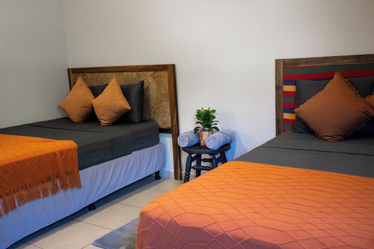 A room with two single beds, each with a wooden headboard, a small table with a plant, and a rolled towel.
