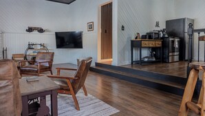 A modern living room with a wooden floor, a dining area with a table and chairs, a kitchen with appliances, and a TV mounted on the wall. - Charming Cabin Hideaway, Peaceful Woodland Escape (Williamston)