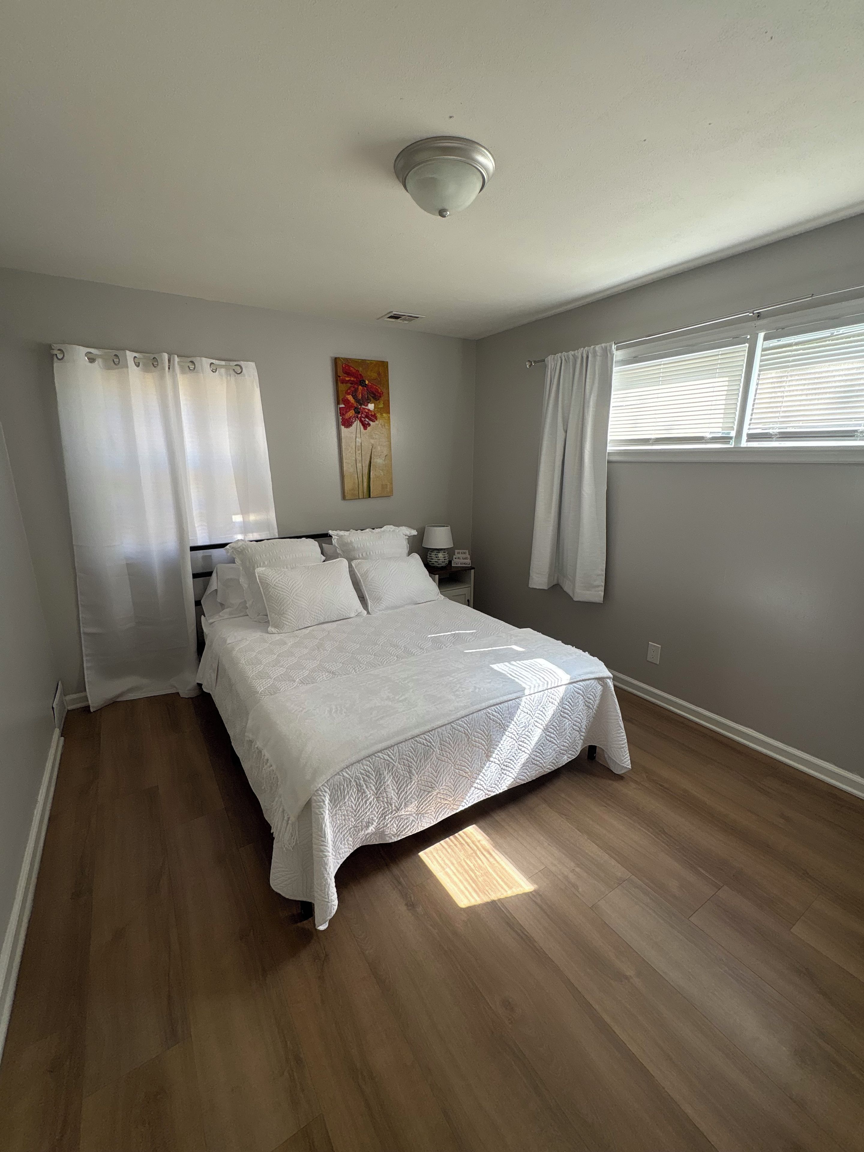 A bedroom with a bed, curtains, a painting, a nightstand, and a window with blinds.