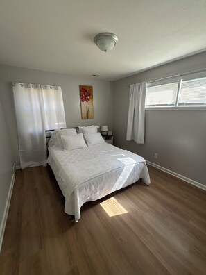 A bedroom with a bed, curtains, a painting, a nightstand, and a window with blinds.