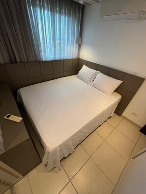 Quarto de hotel com cama grande, mesa de cabeceira, janela com cortinas e ar condicionado.