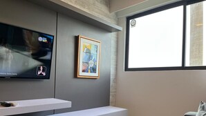 Una sala de estar moderna con una televisión de pantalla plana montada en la pared, un cuadro abstracto enmarcado, un mueble blanco para entretenimiento y un ventanal que proporciona luz natural.