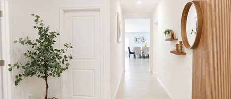 A hallway with a white door, a potted plant, a mirror, and a dining area in the background.