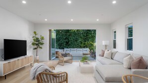 Ein modernes Wohnzimmer mit einem Flachbildfernseher, Korbmöbeln, einem grauen Sofa und Blick auf einen üppigen Grün Garten durch Glasüren.