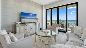 Ein geräumiges Wohnzimmer mit einem großen Glastisch, bequemen Sitzgelegenheiten, einem Fernseher und Blick auf den Strand durch große Fenster.
