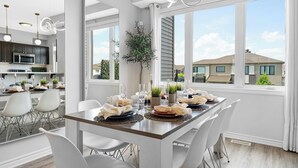 Une cuisine moderne avec des meubles blancs, un coin repas doté d’une longue table dressée pour six personnes, et de grandes fenêtres offrant une vue sur les maisons voisines.