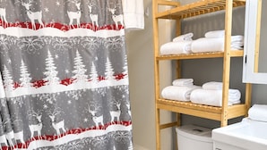Une salle de bain avec un rideau de douche orné d’un motif de forêt hivernale et de cerfs, une étagère en bois avec des serviettes, des toilettes blanches et un meuble blanc.
