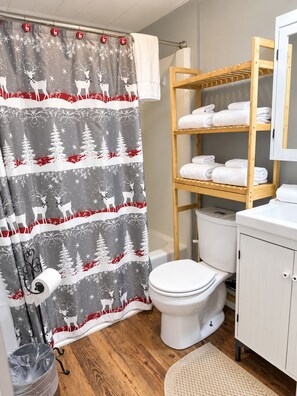 A bathroom with a shower curtain featuring a winter forest and deer design, a wooden shelf with towels, a white toilet, and a white cabinet. - Overlook Condo 21-C Studio ~ Off Mountain ~ 6 miles to Snowshoe Mountaintop Village (Slatyfork)