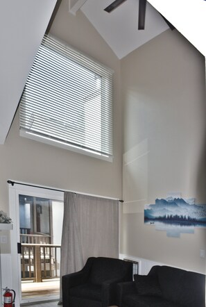 A living room with a sloped ceiling, a large window with blinds, a painting of a mountain landscape, and a fire extinguisher. - Whistlepunk #43: 1 bedroom with Loft, Slopeside (Snowshoe)