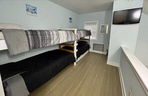 A dormitory room with bunk beds, a television, and a window with blinds.
