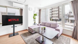 Ein modernes Wohnzimmer mit Flachbildfernseher, einer Couch, einem Couchtisch und einem großen Fenster mit Vorhängen.