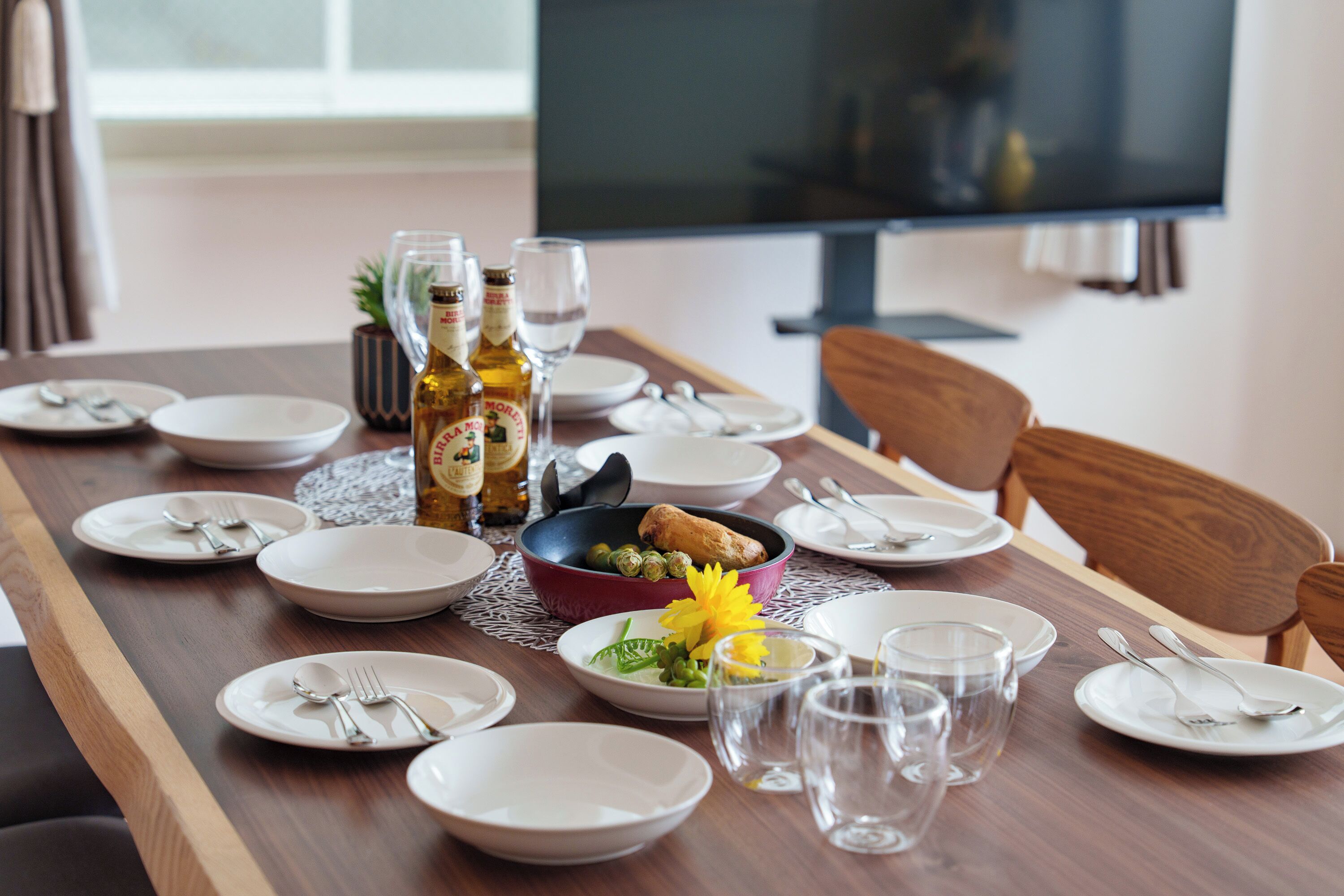 A dining table set with plates, cutlery, glasses, and bottles, with a TV in the background.