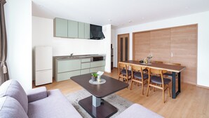 Ein modernes Wohnzimmer mit einem grauen Sofa, einem Esstisch mit vier Stühlen und einer Küchenzeile mit Kühlschrank, Herd und Schränken.
