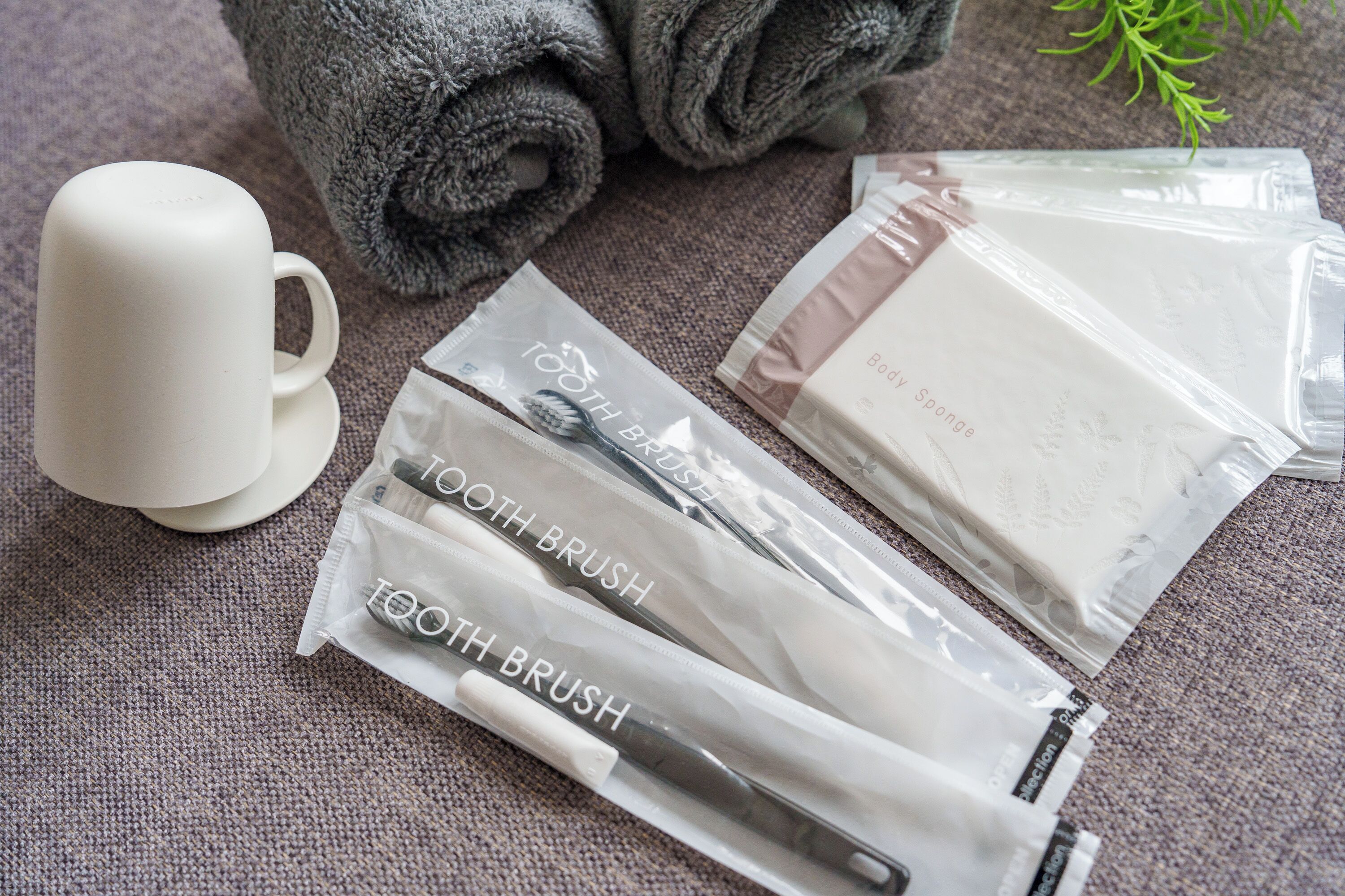 Una taza blanca, dos cepillos de dientes, una pastilla de jabón y una toalla gris enrollada sobre una superficie con textura.