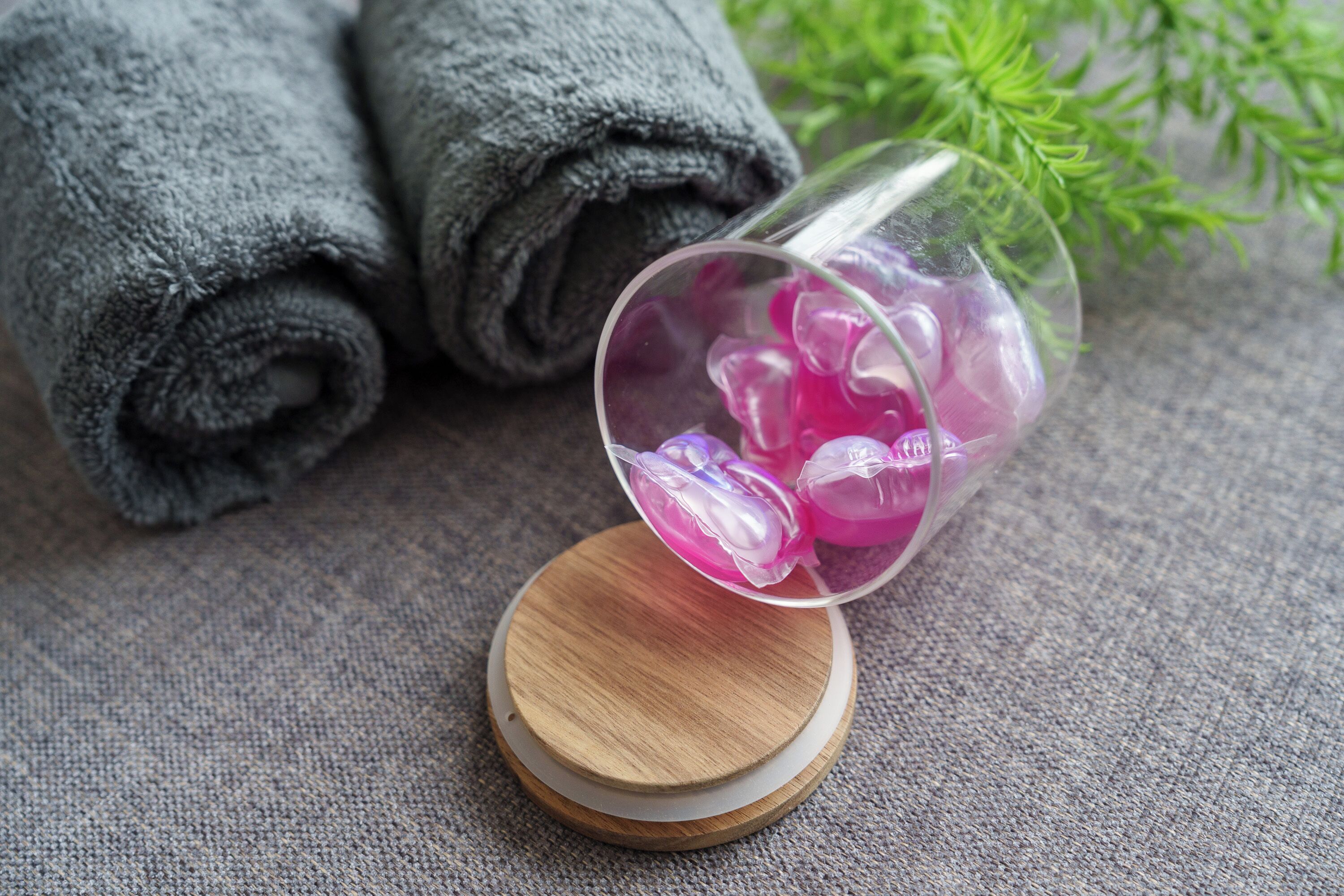A glass with purple bath salts on a wooden coaster, next to rolled gray towels and green plants.
