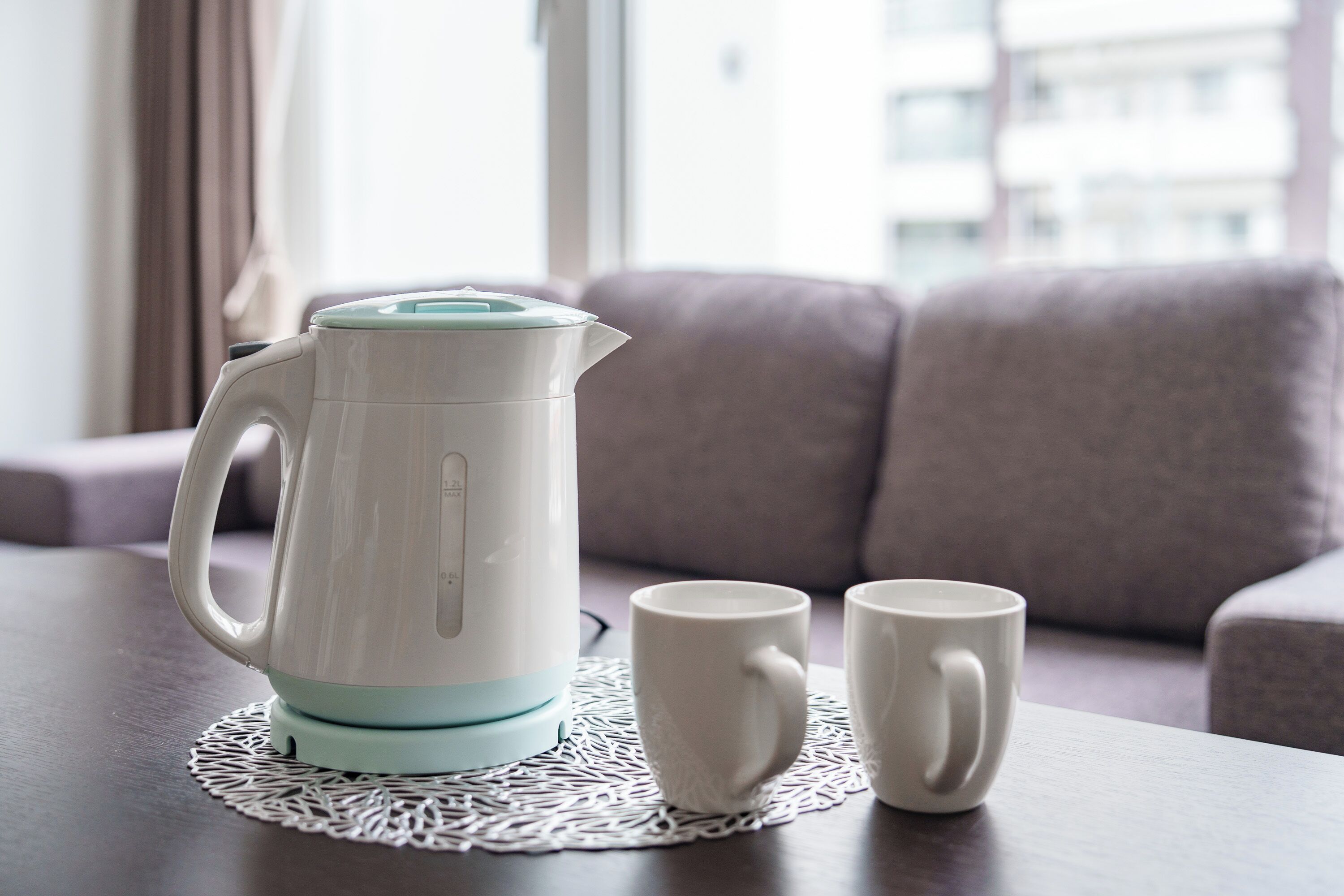 Une bouilloire électrique blanche, dotée d’une base couleur turquoise, est posée sur une table en bois, accompagnée de deux tasses blanches, elles aussi posées sur un chemin de table.
