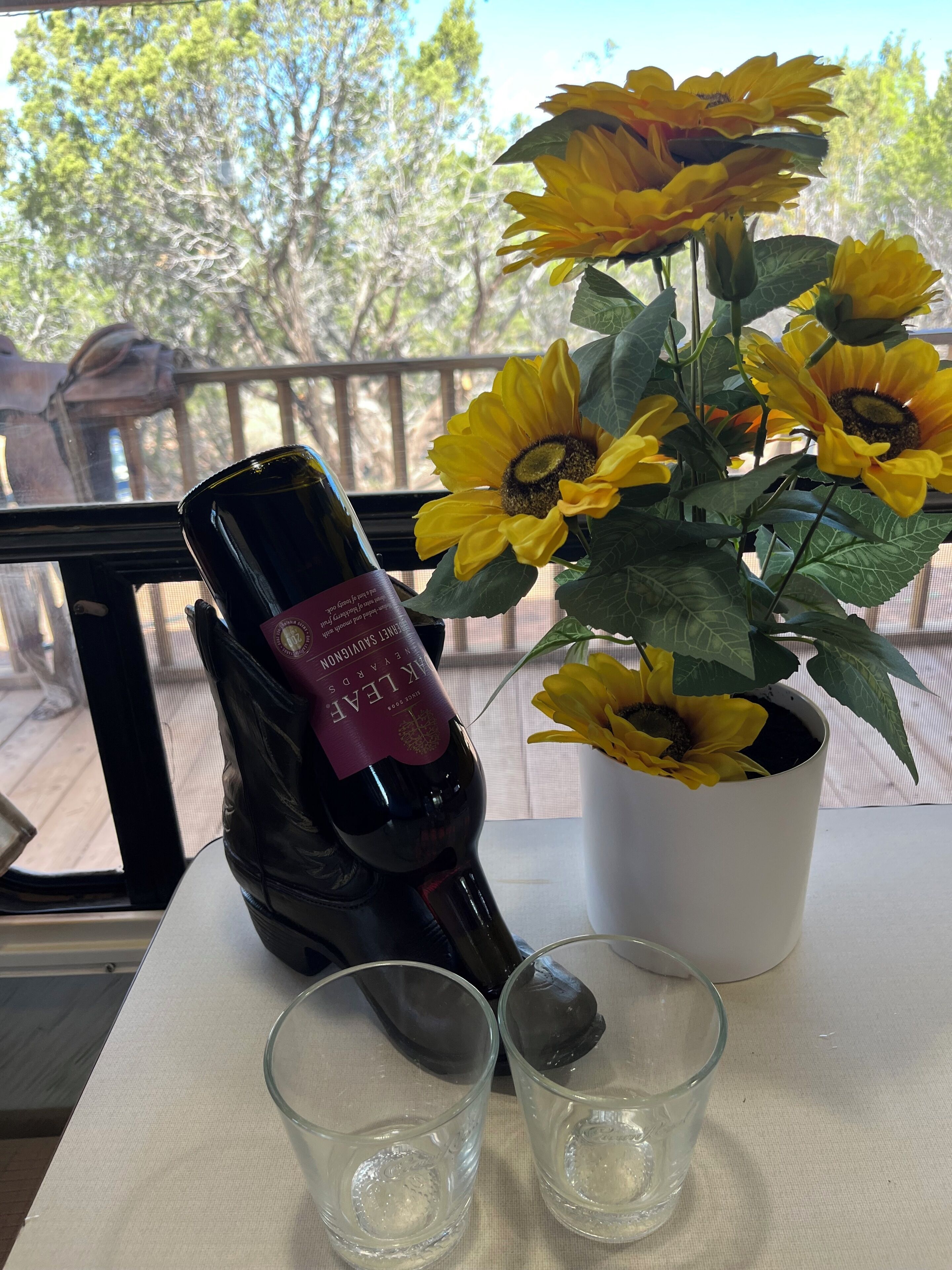 Una botella de vino, dos copas y un girasol en maceta sobre una mesa junto a una ventana.