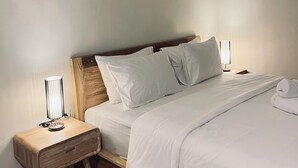 A neatly made bed with white linens, a wooden headboard, and a bedside table with a lamp. - Satria Bali Homestay (Pecatu)