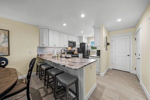A modern kitchen with white cabinets, a granite countertop, and stainless steel appliances. There is a dining area with a wooden table and bar stools. - Gulf Dunes 4 - 2 bed / 2.5 Bath / No Pool / Sleeps 4 - Gulf Front (Boca Grande)