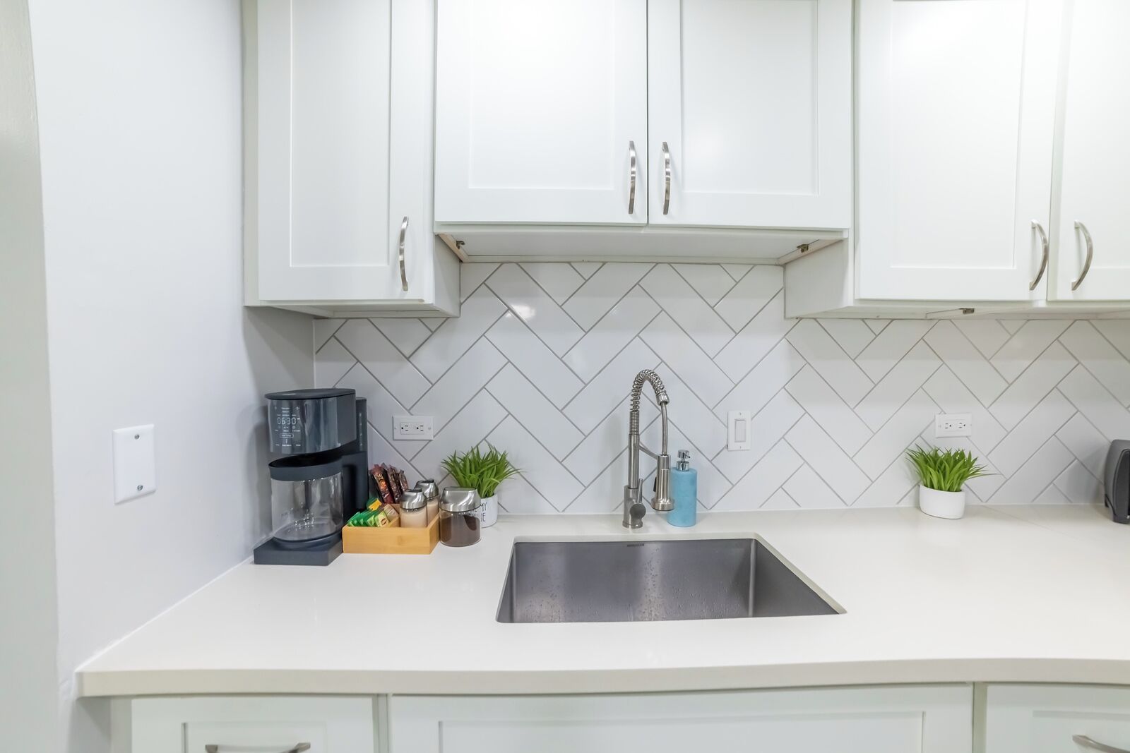 A modern kitchen with white cabinets, a herringbone tile backsplash, a stainless steel sink, a coffee maker, and two potted plants.