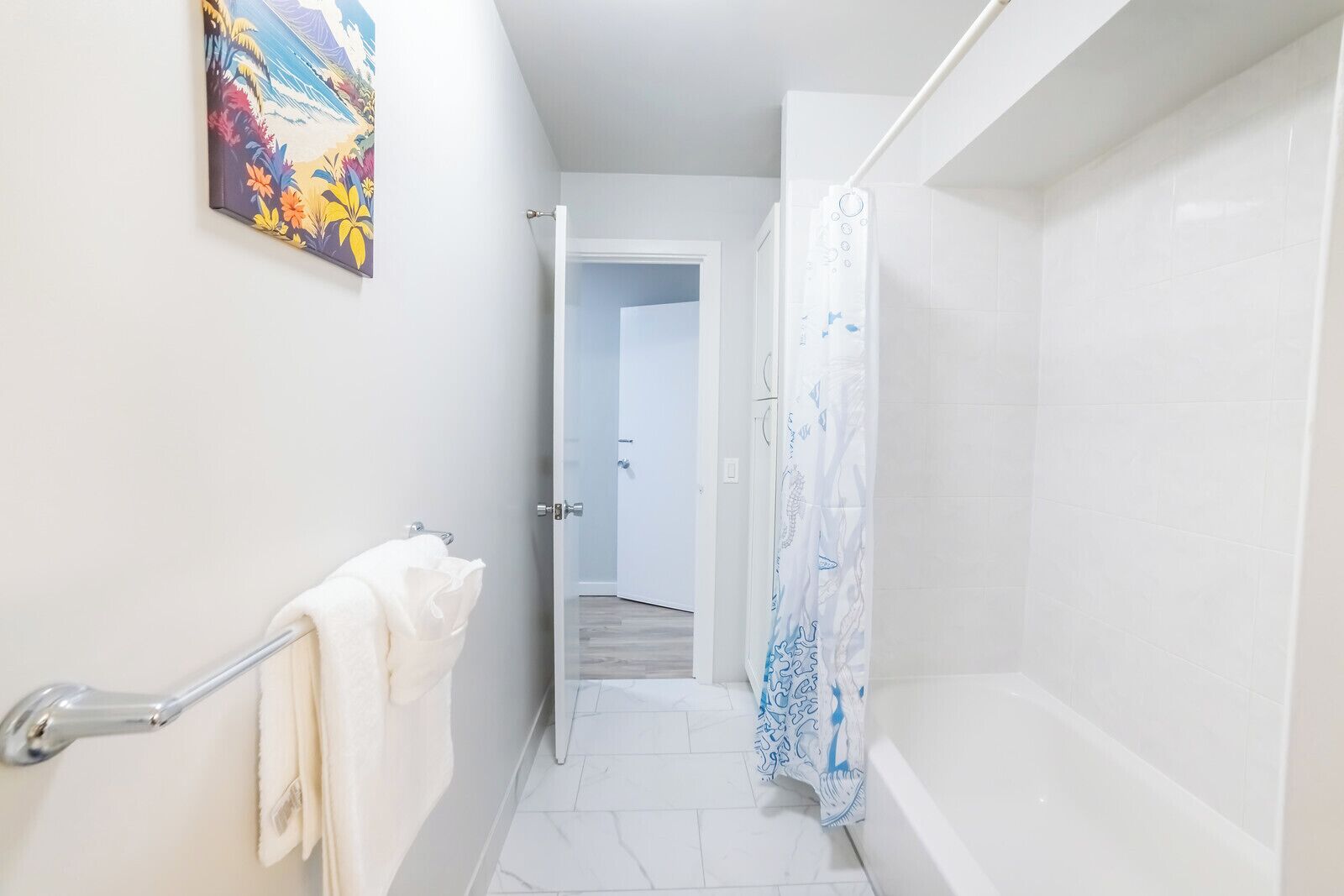 A narrow bathroom with white walls, a shower with a blue and white shower curtain, and a door leading to another room.
