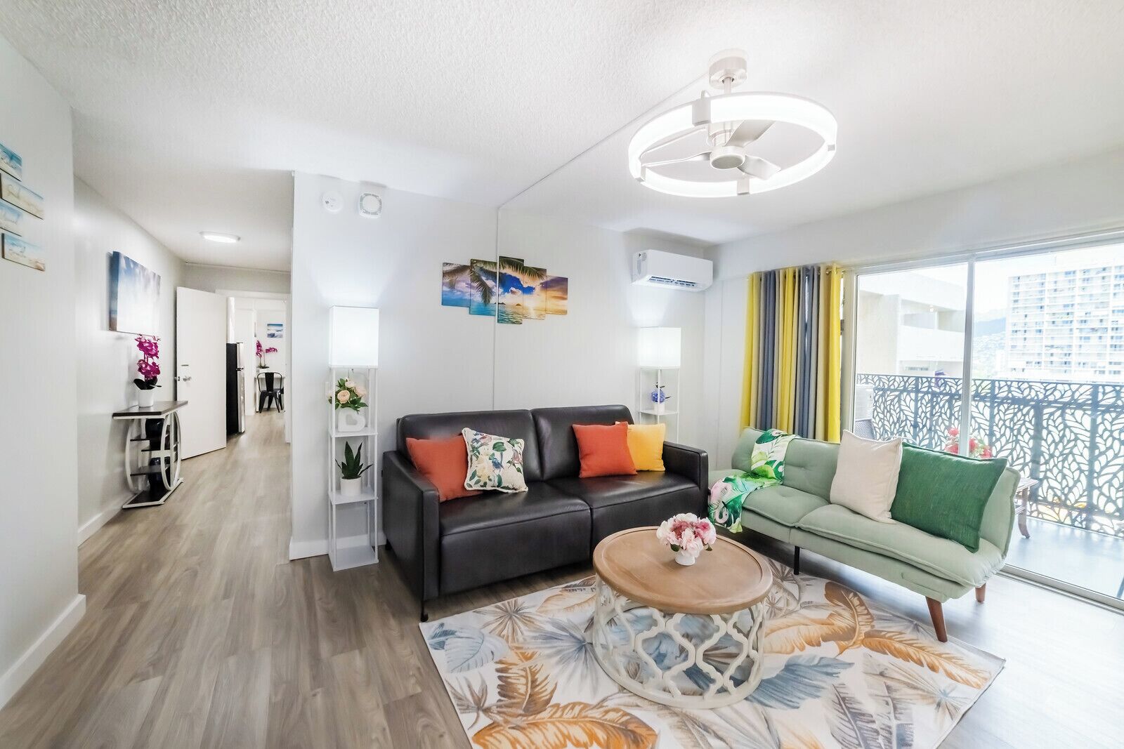 Ein modernes Wohnzimmer mit einer schwarzen Ledersofa, einem grünen Sofa und einem Couchtisch. Auf dem Boden liegt ein Teppich mit floralem Muster. Ein großes Fenster mit gelben Vorhängen und ein Balkon mit einem Metallgeländer sind ebenfalls zu sehen.