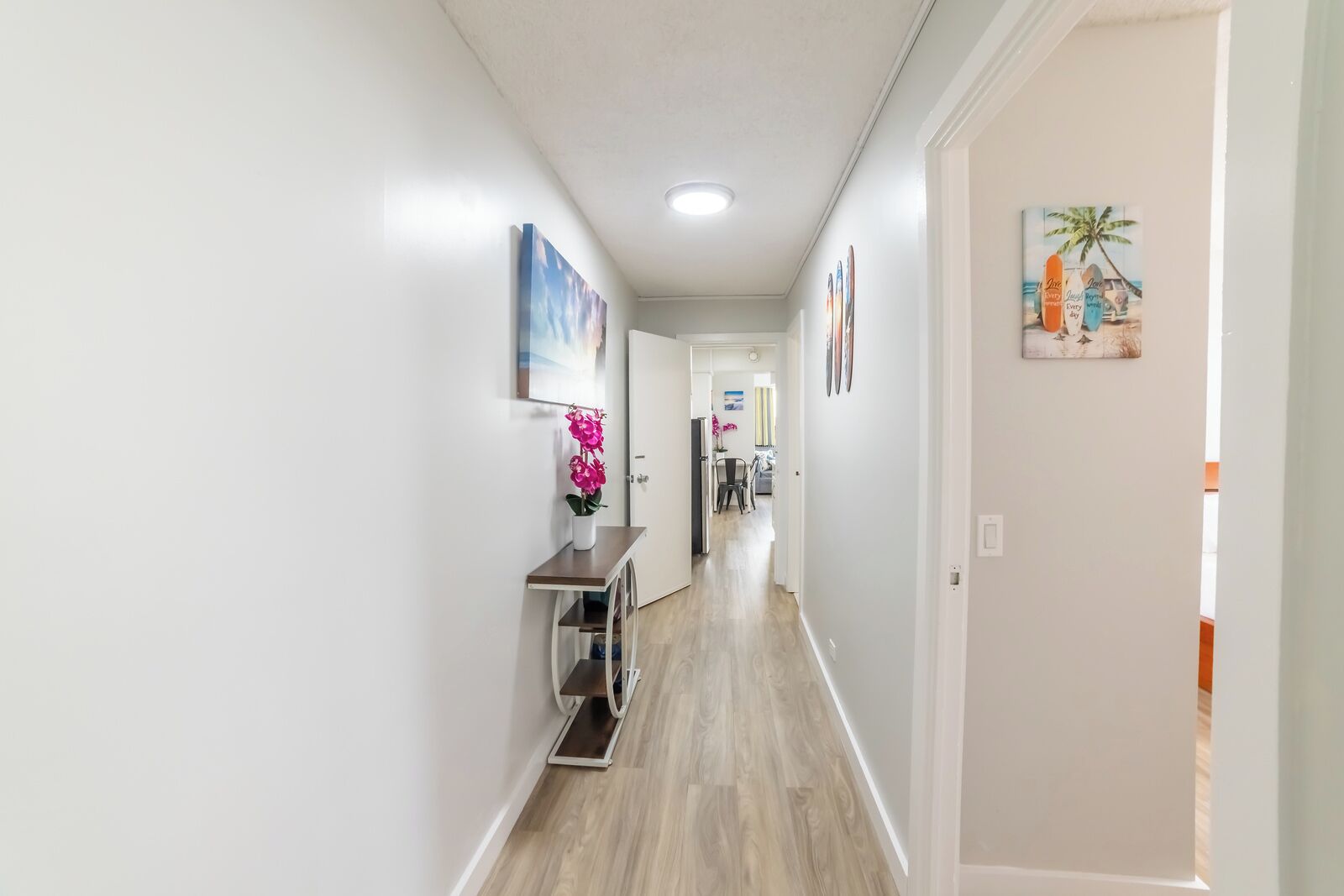 A hallway with a wooden floor, white walls, and a ceiling light. There is a small table with a vase of pink flowers and a framed picture on the wall.