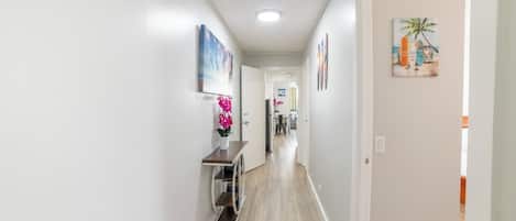 A hallway with a wooden floor, white walls, and a ceiling light. There is a small table with a vase of pink flowers and a framed picture on the wall.