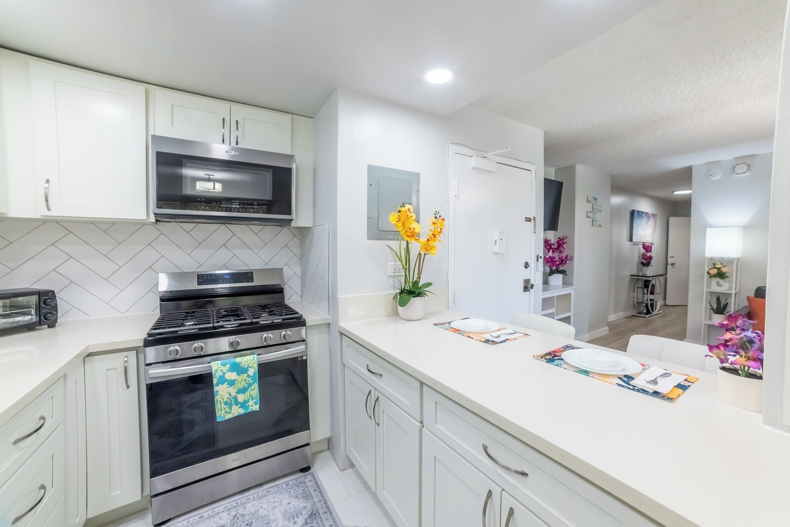 Una cocina moderna con armarios blancos, estufa de acero inoxidable y microondas. Hay una zona de comedor con una mesa dispuesta para dos personas, y una pequeña planta en la encimera.