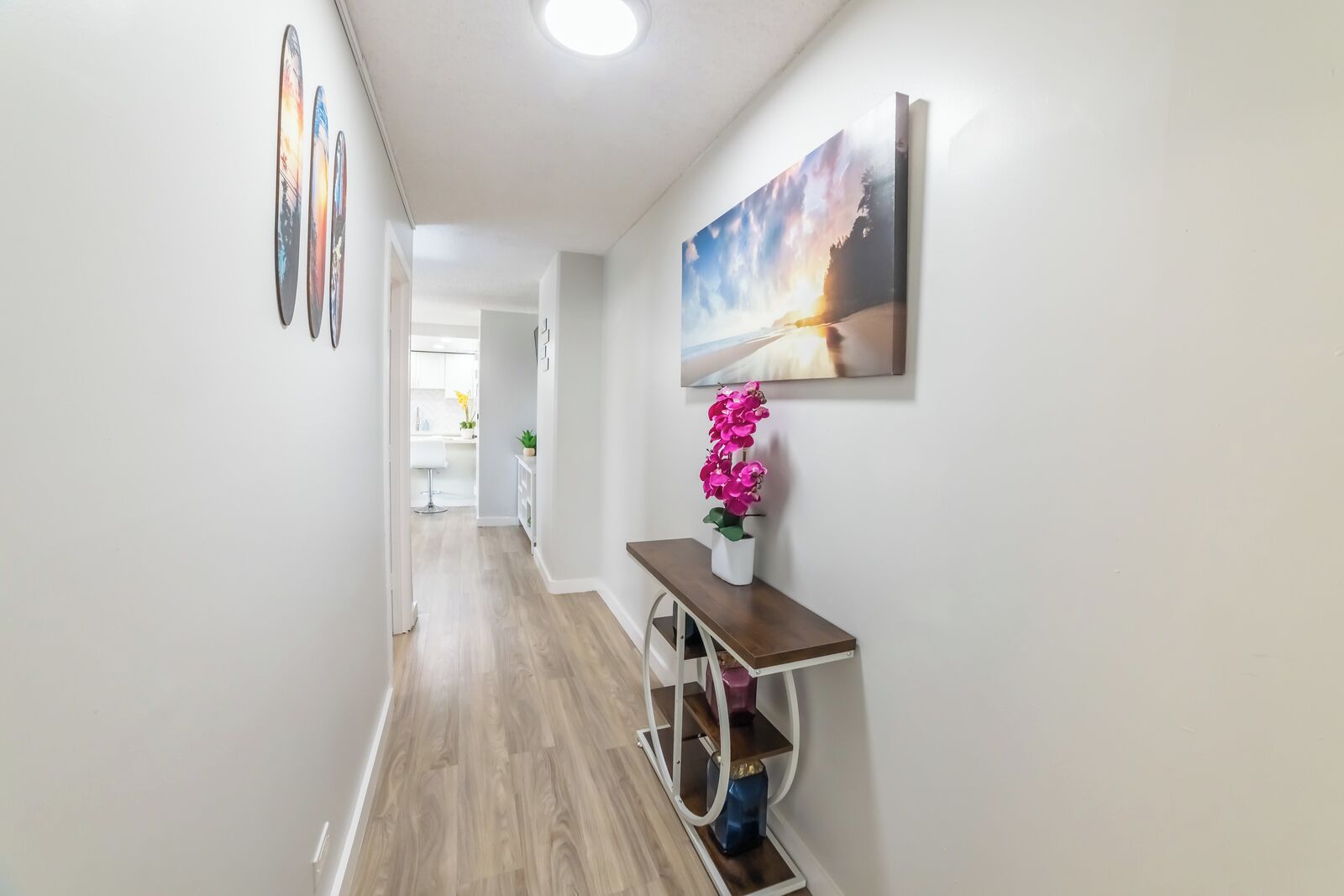 A hallway with a wooden floor, a white wall with a painting, and a small table with a pink flower vase.