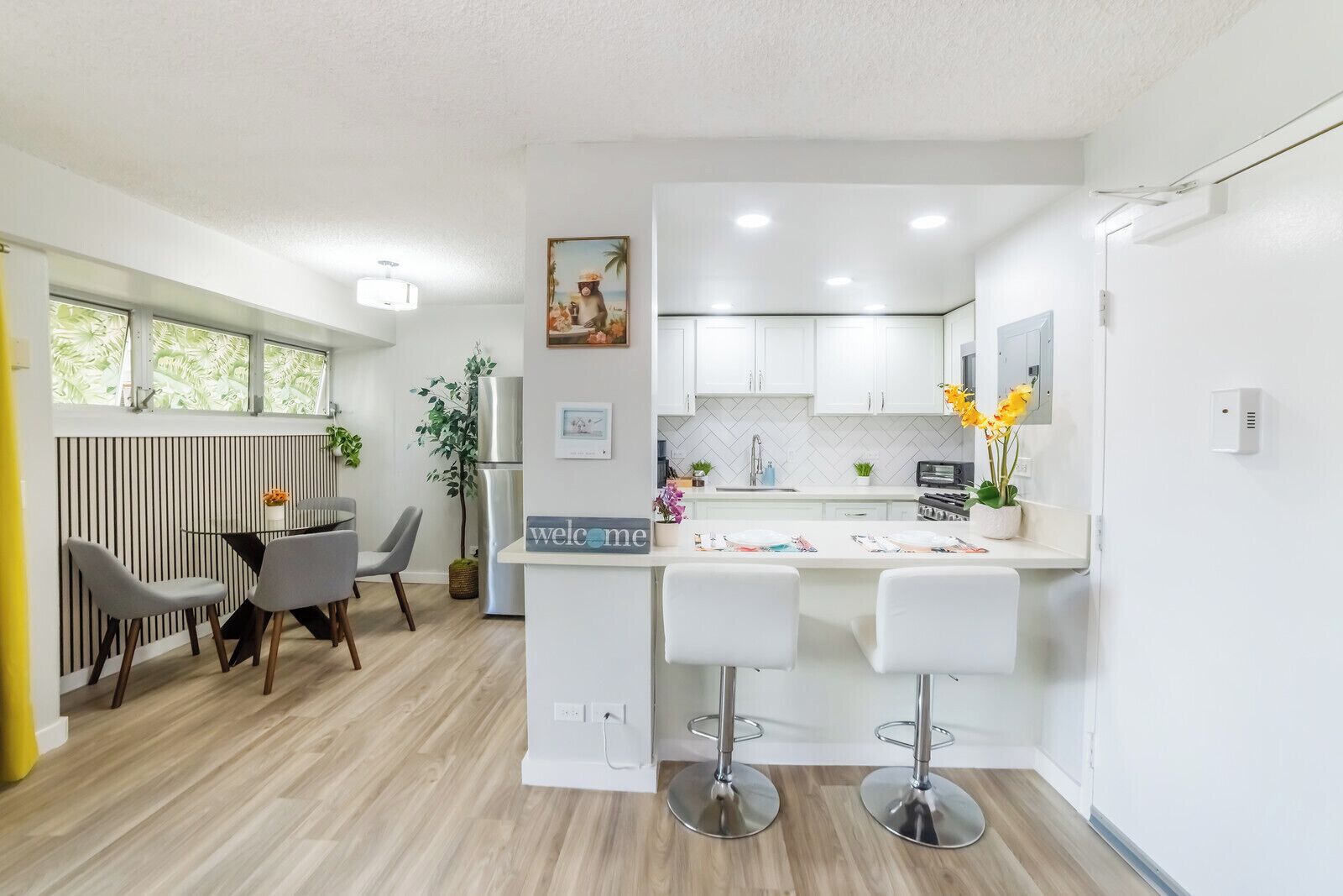 A modern kitchen with white cabinets, a central island with bar stools, a dining area with a table and chairs, and a window with a view of greenery.