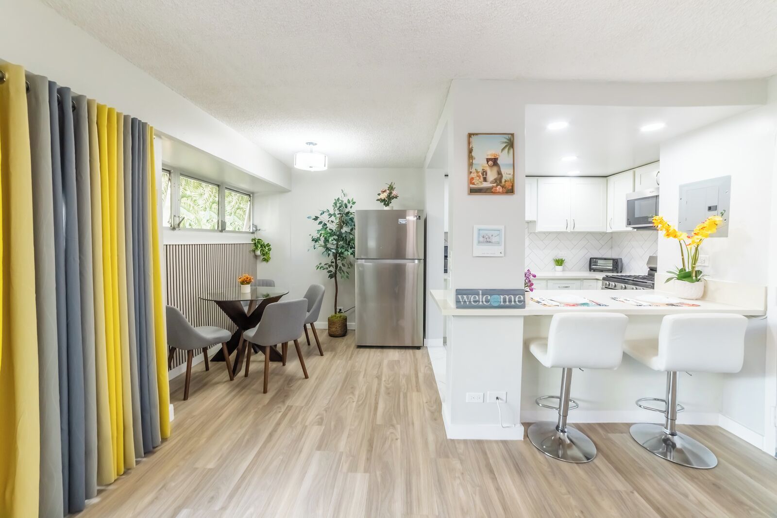 A modern kitchen with a refrigerator, microwave, and stove. There is a dining area with a table and chairs, and a bar area with stools.