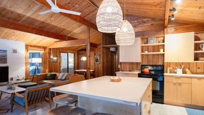 Interior acogedor de una cabaña con techo de madera, una isla central con taburetes, un comedor y una cocina con armarios de madera y una estufa.