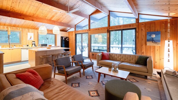 Amplia sala de estar con sofá, sillas y una mesa de centro. Zona de cocina con refrigerador y mesa de comedor. Amplias ventanas ofrecen luz natural y vistas al exterior.