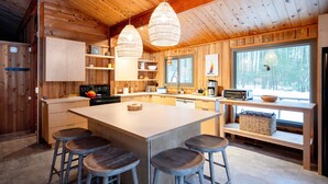 Cocina de cabaña de madera con una isla grande, taburetes de bar, lámparas colgantes y vistas al bosque por la ventana.