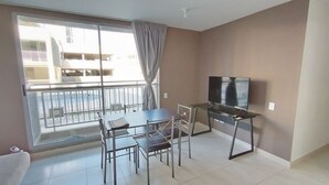 Una stanza con un balcone, una TV su un mobile, un tavolo da pranzo con sedie e un'ampia finestra con tende.