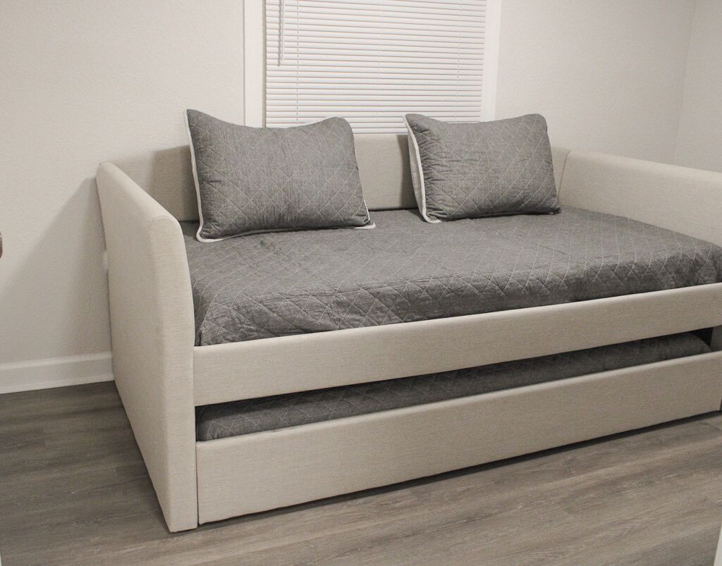 Un sofá moderno con funda gris acolchada y dos almohadones a juego, ubicado frente a una pared blanca con un ventanal.