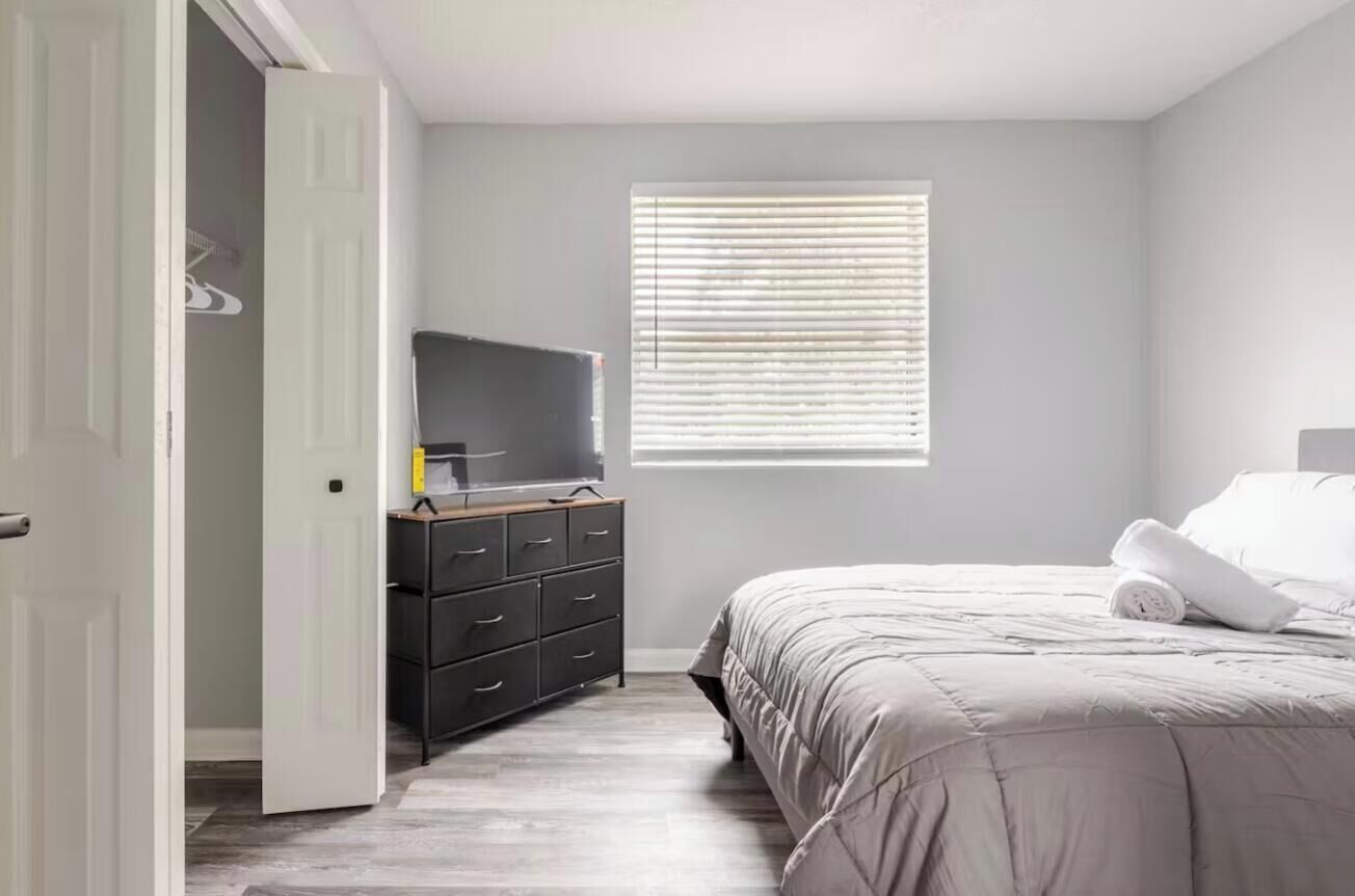 A bedroom with a bed, a dresser, a television, and a window with blinds.