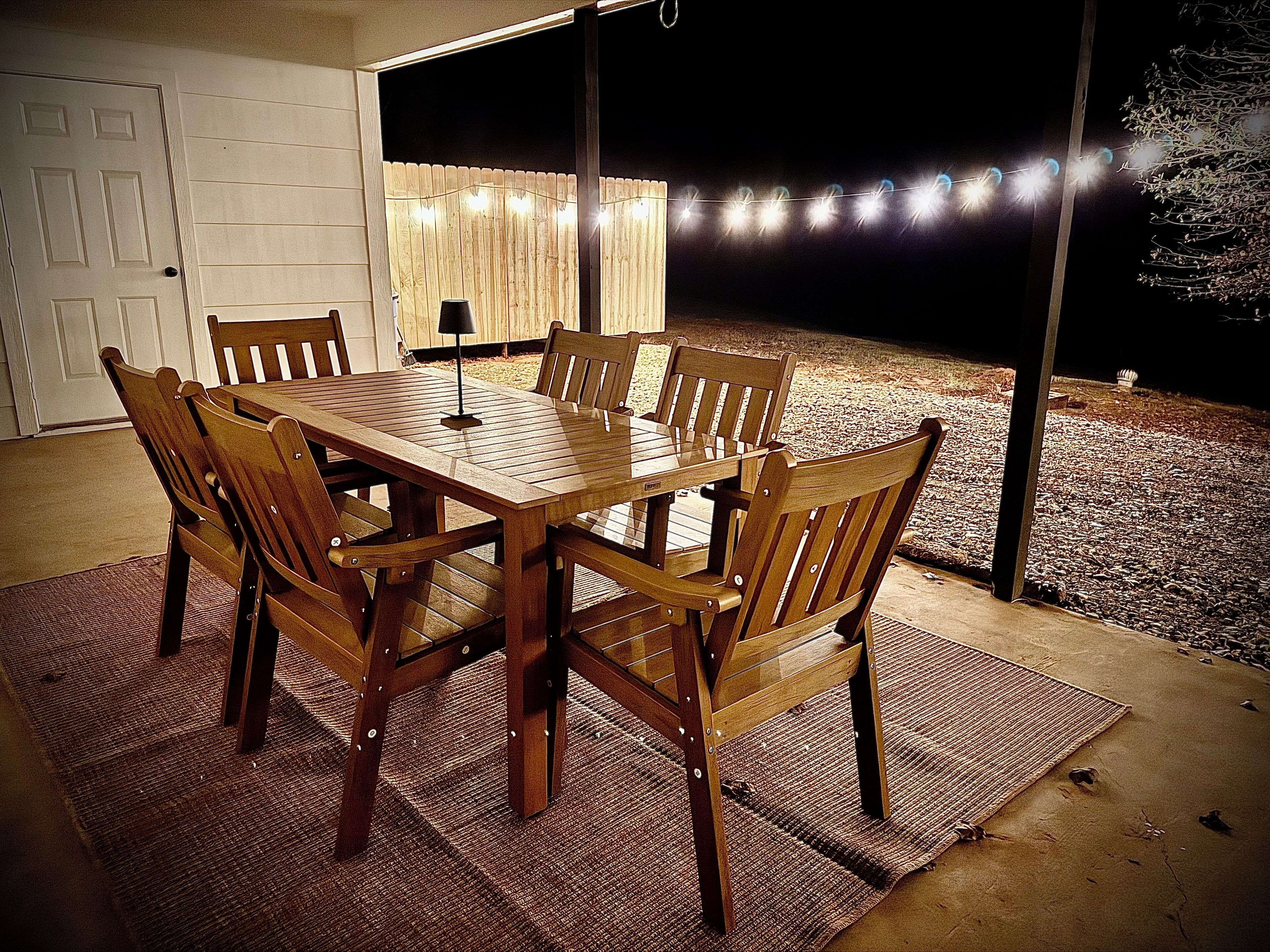 Een buiten eethoek met een houten tafel en stoelen, een vloerkleed en sfeerverlichting.