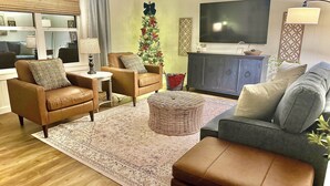 A living room with a brown leather sofa, a grey couch, a small round ottoman, a large flat-screen TV, and a Christmas tree.