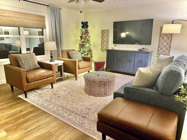 A living room with a brown leather sofa, a grey couch, a small round ottoman, a large flat-screen TV, and a Christmas tree.