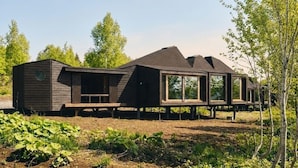 Une cabane moderne en bois, dotée de grandes fenêtres et d’un toit en pente, située dans un environnement naturel, au milieu des arbres et de la verdure.