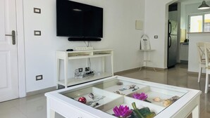 Un salon moderne avec un téléviseur à écran plat fixé au mur, une console blanche sur laquelle est posé un panneau, une table basse en verre agrémentée d’objets décoratifs, et un coin cuisine visible à travers une porte ouverte.