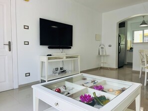 Ein modernes Wohnzimmer mit einem an der Wand befestigten Flachbildfernseher, einem weißen Konsolentisch mit einem Schild, einem Glastisch mit Dekoration und einem durch einen offenen Durchgang einsehbaren Küchenbereich.