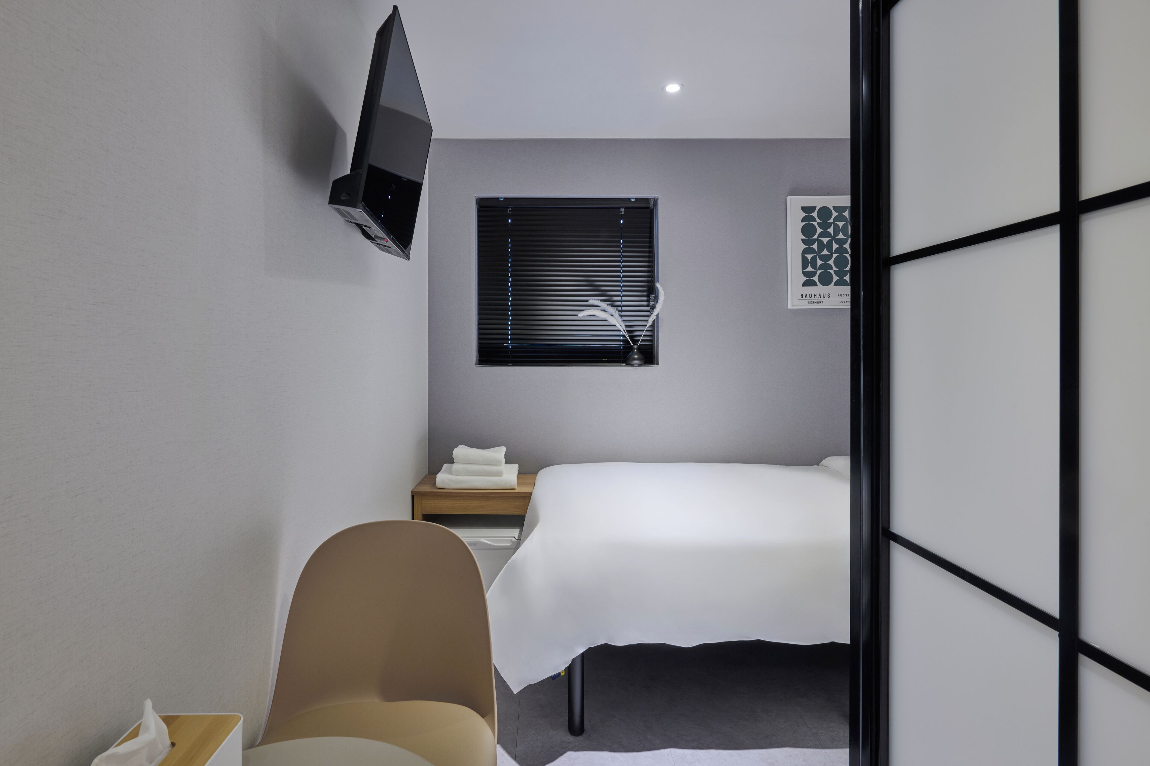 A modern hotel room with a bed, a bedside table with a lamp, a chair, and a TV mounted on the wall.