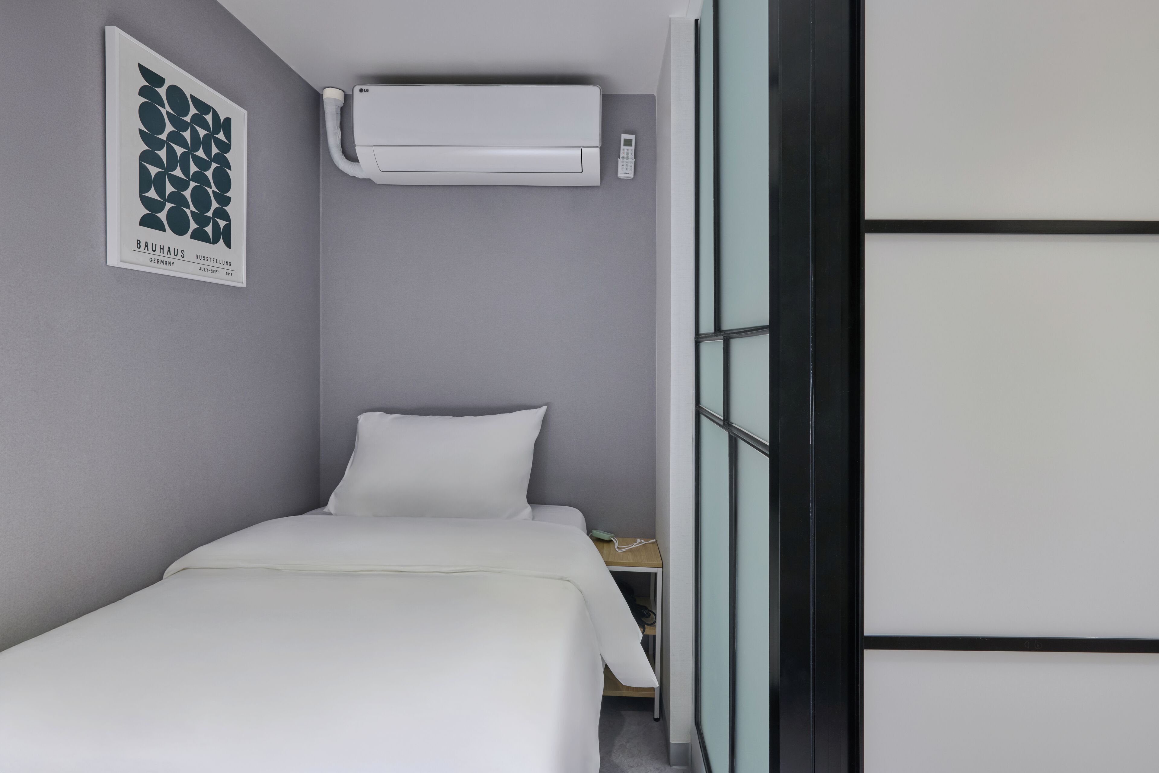 A modern hotel room with a bed, a wall-mounted air conditioner, a small table with a telephone, and a framed picture on the wall.