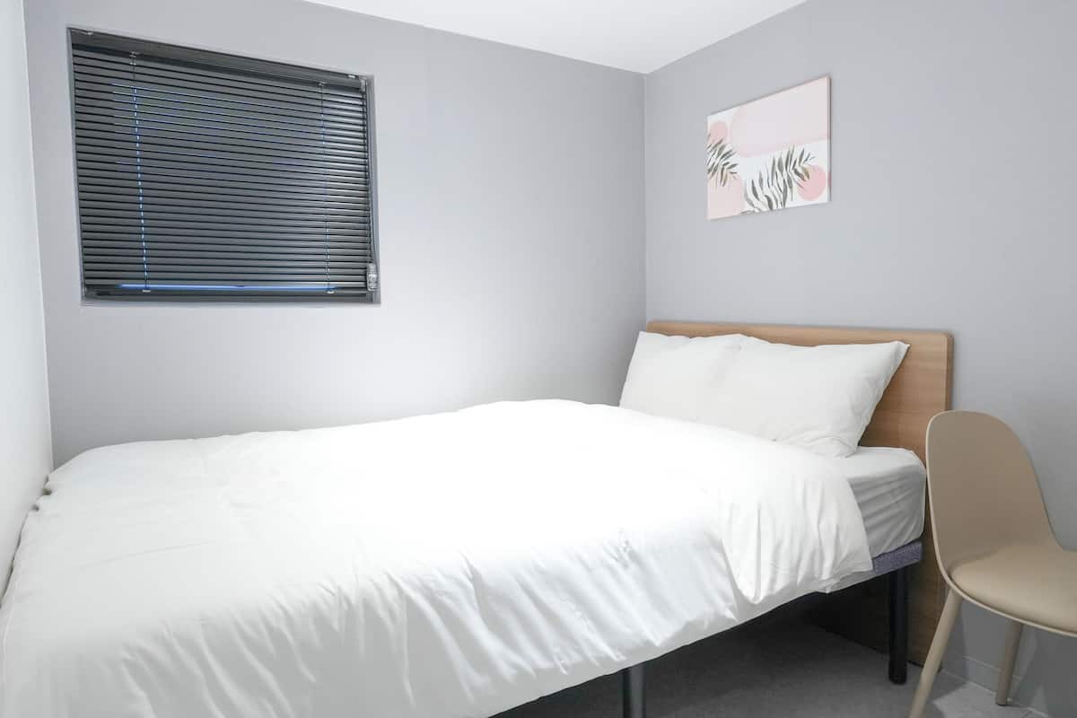 Un dormitorio con cama, una silla, una mesita, una ventana con persianas y un cuadro colgado en la pared.
