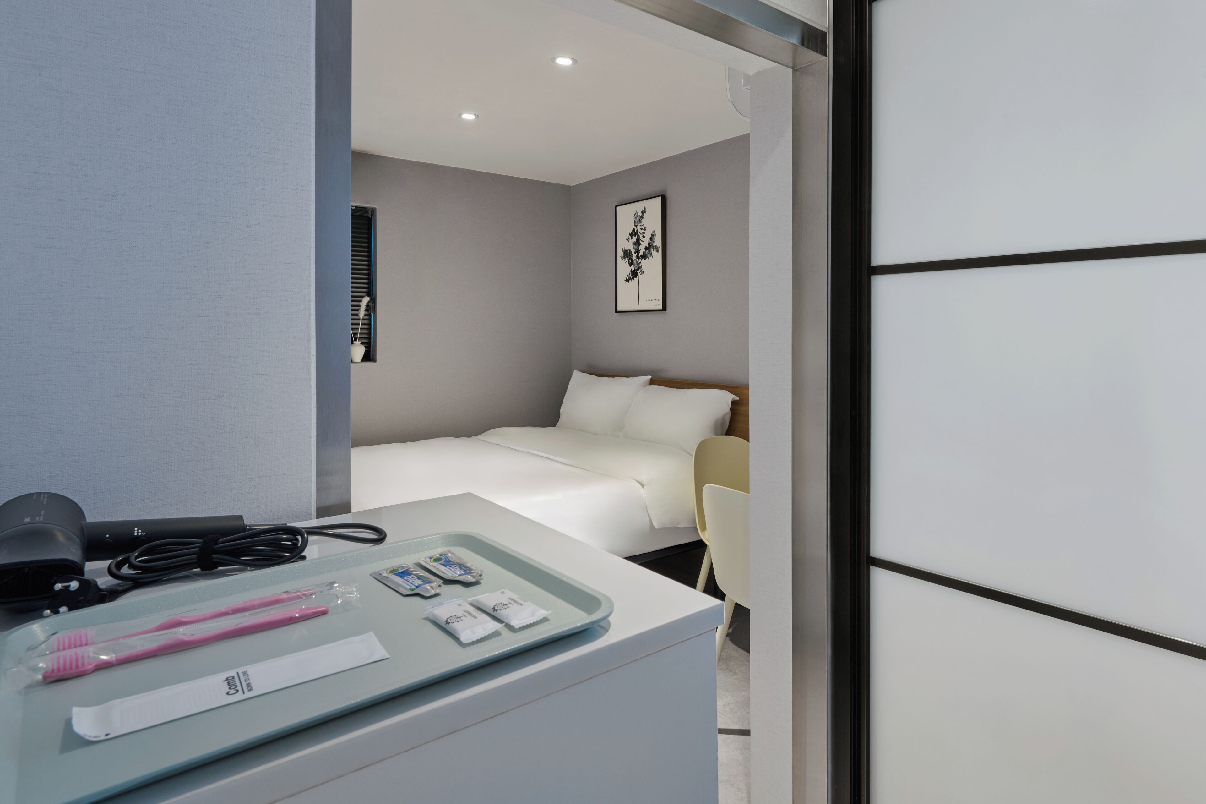 A hotel room with a bed, bedside table, and a chair. There is a framed picture on the wall and a window with blinds.