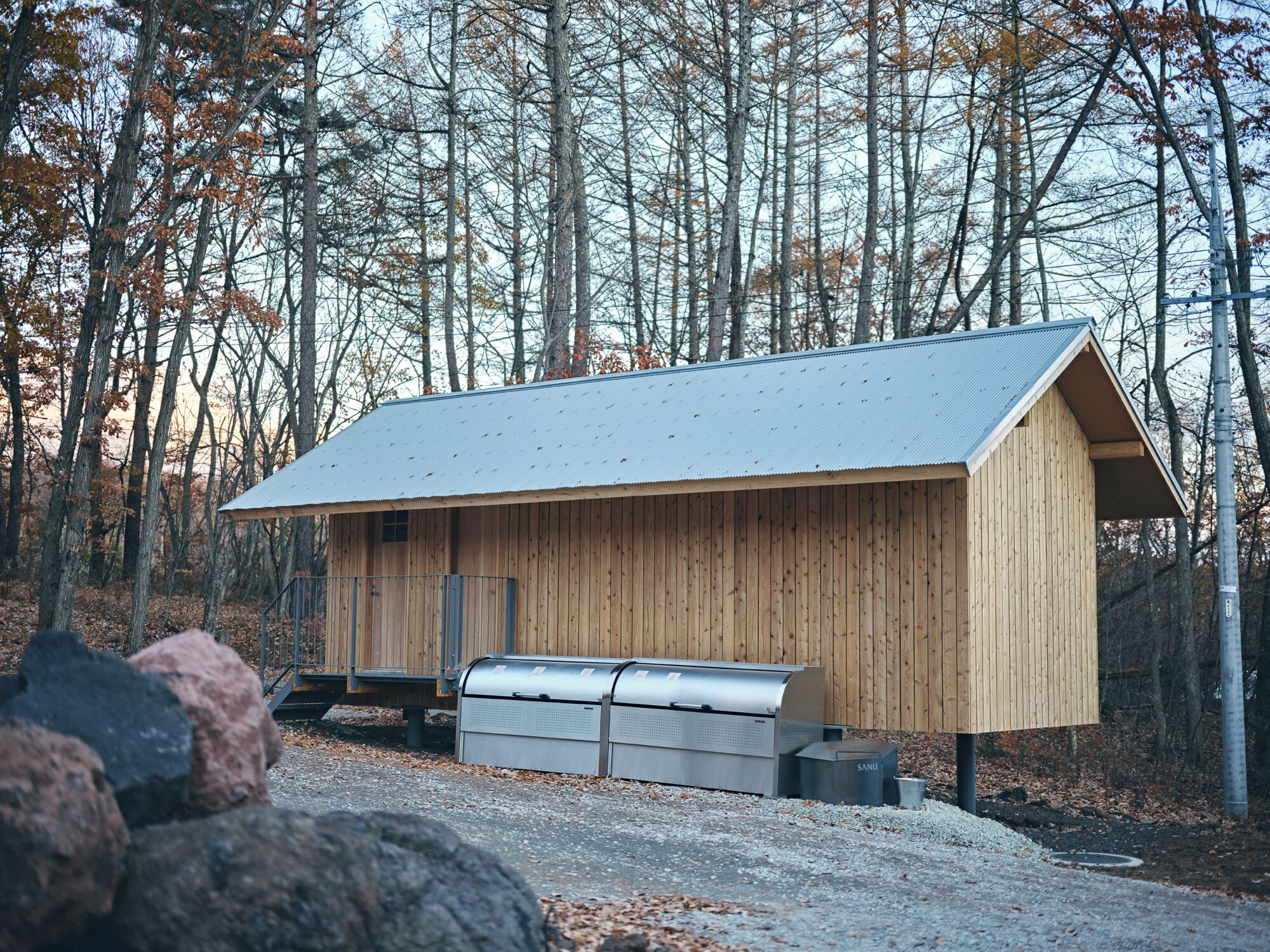 A wooden cabin with a metal roof, a metal bench, and a trash can labeled "SANU" in a forested area.